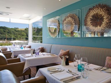 A dining area with tables set with white tablecloths glasses and cutlery by a window with a view of trees and water at 21 Dart Marina in Dartmouth