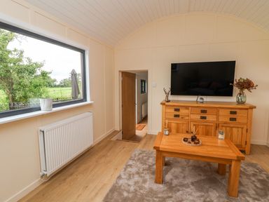 A room with a wooden table and cabinet a wall mounted TV and a window showing outdoor greenery at The View in Bangor-On-Dee