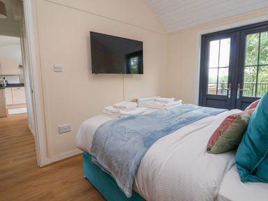 A bedroom with a bed, pillows, folded towels, a wall-mounted television, wooden floor, and glass doors at The View in Bangor-On-Dee