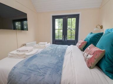 A bedroom with a bed covered with blue blanket and teal pillows with pink decorative cushions near double glass doors and a wall-mounted television at The View in Bangor-On-Dee