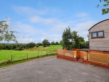 A gravel driveway next to a wooden building with a fenced patio and a grassy field with trees in the background at The View in Bangor-On-Dee