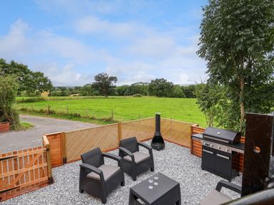 An outdoor patio with black wicker chairs a small table a chiminea a barbecue grill and a wooden fence overlooking green fields at The View in Bangor-On-Dee