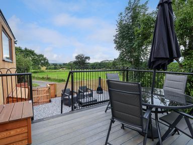 An outdoor balcony with a glass table chairs and a closed umbrella overlooking a garden with a seating area and green fields at The View in Bangor-On-Dee