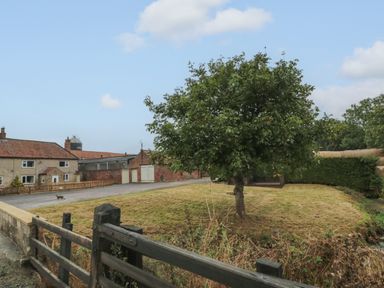 A grassy area with a tree and a wooden fence near stone and brick buildings at Bellmanear Farm Cottage in North Grimston near Malton