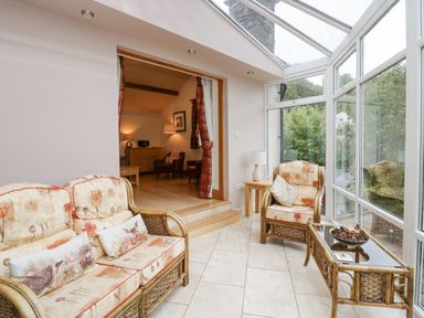 A sunroom with wicker sofa and armchair and glass coffee table next to large windows and a door leading to a living room at Blue Hill House in Ambleside