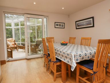 A dining room with a table covered by a patterned tablecloth and six wooden chairs next to glass doors leading to a sunroom with wicker furniture