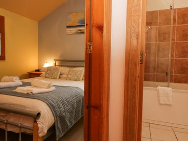 A bedroom with a bed covered with a gray quilt and rolled towels next to a bathroom with a bathtub and tiled walls at Blue Hill House in Ambleside
