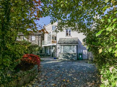 A white house with stone and glass features surrounded by trees and a gravel driveway at Blue Hill House in Ambleside