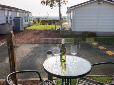 A patio table with a bottle and two glasses between two chairs with views of a driveway and houses at Brocklebank Lodge 35 High Hesket near Armathwaite