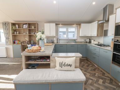 A kitchen with blue and white cabinets a built in oven a gas stove a window above the sink a cushioned bench with a welcome pillow and shelves with books Brocklebank Lodge 35 High Hesket near Armathwaite