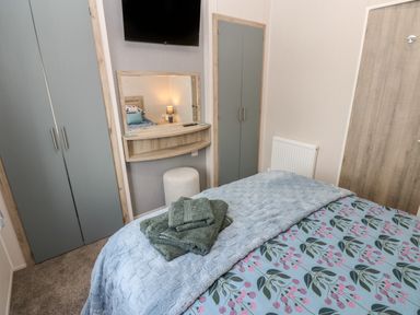 A bedroom with a bed covered in a blue and floral bedspread towels on the bed a wall-mounted television a mirror with a small shelf and a stool two wardrobes and a door Brocklebank Lodge 35 High Hesket near Armathwaite