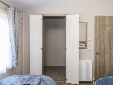 A bedroom with an open wardrobe, a wooden door, a radiator, a window with patterned curtains, and two beds with blue bedding at Brocklebank Lodge 35 High Hesket near Armathwaite