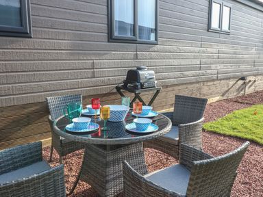 An outdoor patio dining set with four chairs a round glass table set with colorful plates bowls and glasses beside a wall with windows and a barbecue grill at Brocklebank Lodge 35 in High Hesket near Armathwaite