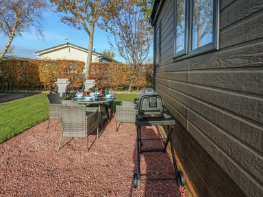 An outdoor patio with a glass table set with dishes and chairs next to a barbecue grill by a wooden house at Brocklebank Lodge 35 in High Hesket near Armathwaite