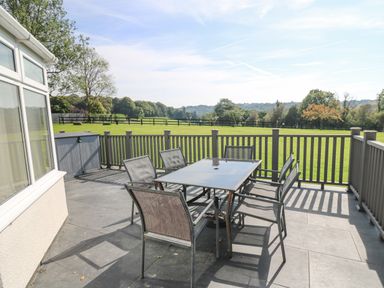 An outdoor patio with a glass table and six chairs overlooking a fenced green field at Trem-Yr-Afon in Aberaeron