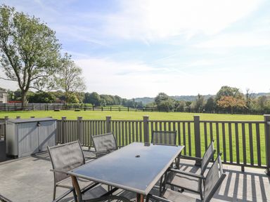 An outdoor patio with a glass table and six chairs overlooking a fenced grassy field with trees in the background at Trem-Yr-Afon in Aberaeron