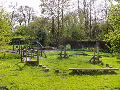 An outdoor playground with wooden climbing structures slides and stepping logs in a grassy area surrounded by trees at Trem-Yr-Afon in Aberaeron