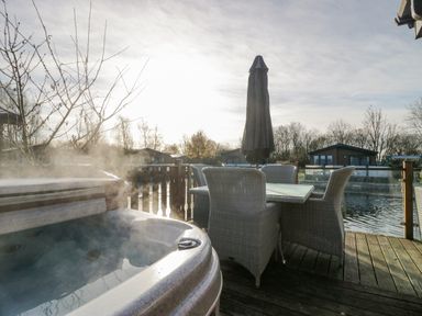 An outdoor seating area with a table and four wicker chairs next to a steaming hot tub on a wooden deck by the water at The Three Bears Lodge Coniston Drive 11 South Lakeland Leisure Village near Warton