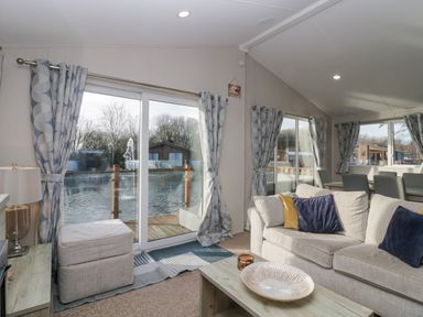 A living room with a beige sofa and blue and yellow cushions next to large windows with patterned curtains at The Three Bears Lodge Coniston Drive 11 South Lakeland Leisure Village near Warton