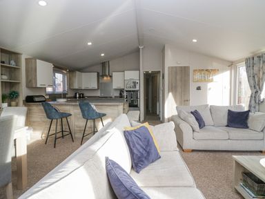 An open living room and kitchen with sofas, stools at a kitchen island, and a dining table at The Three Bears Lodge, Coniston Drive 11 South Lakeland Leisure Village near Warton