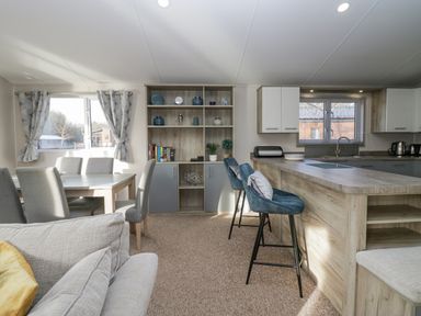 A dining area with a table and six chairs next to a living room with a sofa and cushions and a kitchen with a countertop and two bar stools at The Three Bears Lodge Coniston Drive 11 South Lakeland Leisure Village near Warton