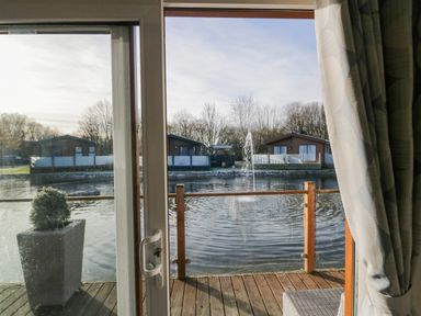 A view of a pond with a water fountain and wooden cabins across the water seen through a glass door with curtains at The Three Bears Lodge Coniston Drive 11 South Lakeland Leisure Village near Warton