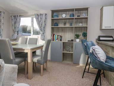 A dining area with a wooden table and six grey chairs next to window with curtains and a shelving unit with books plants and decorative items at The Three Bears Lodge Coniston Drive 11 South Lakeland Leisure Village near Warton