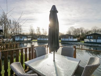 An outdoor patio with a table, umbrella, and four chairs overlooking a water body and several wooden cabins at The Three Bears Lodge, Coniston Drive 11, South Lakeland Leisure Village near Warton
