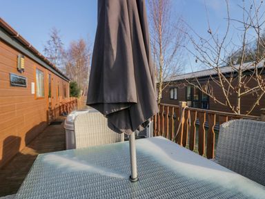 An outdoor patio table with a closed umbrella and chairs near wooden lodges and trees at The Three Bears Lodge Coniston Drive 11 South Lakeland Leisure Village near Warton