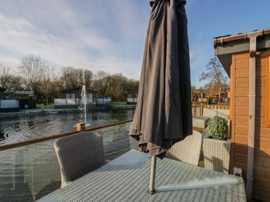 An outdoor patio with wicker chairs a glass table and a closed umbrella overlooking a water feature at The Three Bears Lodge Coniston Drive 11 South Lakeland Leisure Village near Warton
