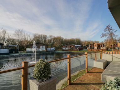 A deck with outdoor seating and a planter overlooking a pond with a fountain and several small buildings at The Three Bears Lodge Coniston Drive 11 South Lakeland Leisure Village near Warton