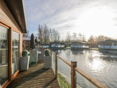 A wooden deck with wicker chairs and a table next to a water body with houses in the background at The Three Bears Lodge Coniston Drive 11 South Lakeland Leisure Village near Warton