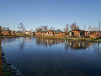 A frozen pond with wooden lodges and trees in the background at The Three Bears Lodge Coniston Drive 11 South Lakeland Leisure Village near Warton
