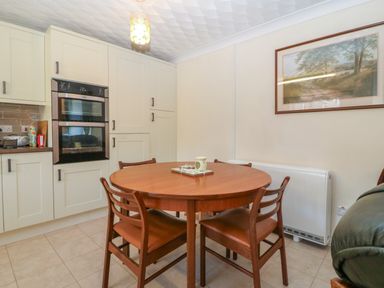 A kitchen with a round wooden dining table and four chairs a double oven and wall cabinets with a landscape painting on the wall at Gwynlys in Cemaes Bay