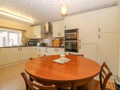 A kitchen with a round wooden table and chairs with white cabinets and built in oven at Gwynlys in Cemaes Bay