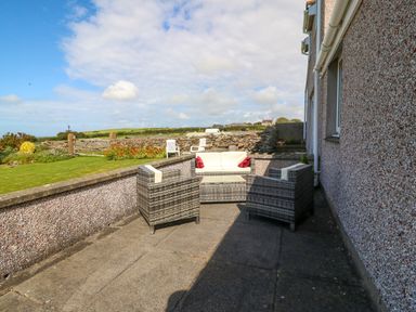 An outdoor patio with wicker chairs and a sofa overlooking a garden at Gwynlys in Cemaes Bay