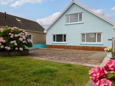 A blue house with three windows a stone driveway and flowers in the garden at Carousel in Broad Haven