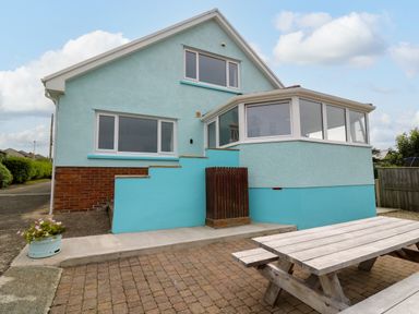 The exterior of a blue house with a brick lower section a wooden picnic table on a paved patio and a small flower pot near the driveway at Carousel in Broad Haven