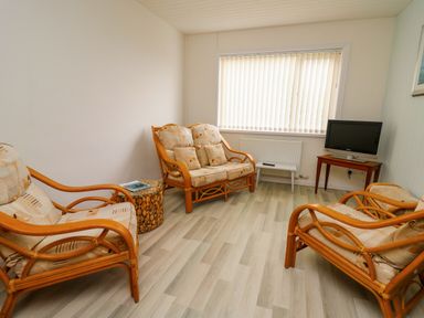 A small living room with three wooden framed cushioned chairs a small side table a television on a wooden stand and a window with vertical blinds at Carousel in Broad Haven