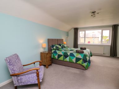 A bedroom with a double bed with geometric bedding a wooden bedside table with a lamp a purple armchair and large window at Carousel in Broad Haven