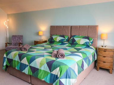 A bedroom with a double bed with green and blue geometric bedspread and pillows two bedside tables with lamps and a chair with rolled towels at Carousel Broad Haven