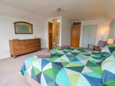A bedroom with a bed covered in a geometric green and blue duvet a wooden dresser with a mirror and an armchair at Carousel in Broad Haven