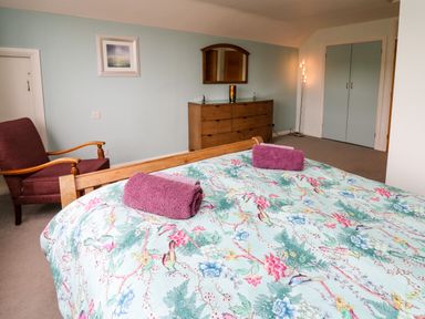 A bedroom with a bed with floral bedding and two purple towels a wooden dresser with a mirror a purple armchair and a floor lamp at Carousel in Broad Haven
