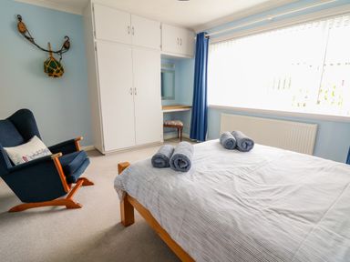 A bedroom with a bed with rolled towels a blue armchair a white wardrobe and a window with blue curtains at Carousel in Broad Haven