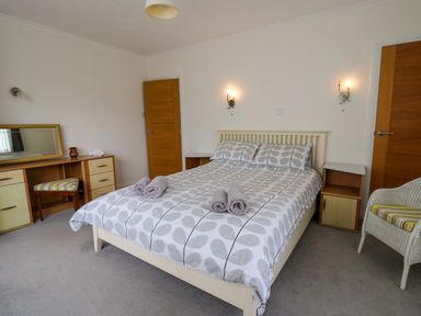 A bedroom with a double bed with patterned bedding two rolled towels dressing table with mirror wooden doors and a chair at Carousel in Broad Haven