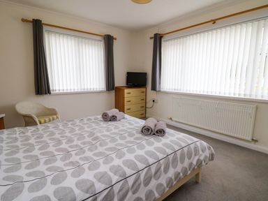 A bedroom with a bed with rolled towels on it a chair a chest of drawers with a small television and two windows with curtains at Carousel in Broad Haven