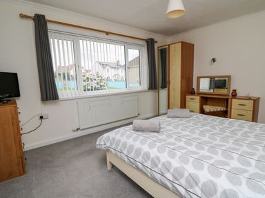 A bedroom with a bed, wardrobe, dressing table, television, and window with vertical blinds at Carousel in Broad Haven