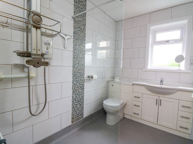 A bathroom with a toilet sink with storage cabinets a walk in shower with wall tiles and a window at Carousel in Broad Haven