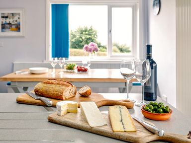 A kitchen countertop with bread cheese olives wine and wine glasses with a dining table and window in the background at Carousel in Broad Haven