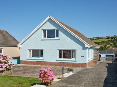 A light blue house with a sloped roof and large windows with pink flowers in front at Carousel in Broad Haven